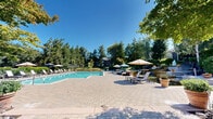 Located at 7300 Parkwood Cir in Dublin, California, this expansive outdoor amenity area is designed to provide a resort-style recreational experience. At the heart of the space is a generously sized swimming pool with crystal-clear turquoise water, surrounded by a well-maintained paver and wood-deck pool surround. Rows of lounge chairs and umbrella-shaded seating are arranged along the poolside, offering ample space for sunbathing and relaxation under the Northern California sun. A separate spa hot tub is situated adjacent to the main pool, tucked within a neatly landscaped enclosure with hedging for added privacy. The entire area is framed by towering mature trees — including redwoods, birches, and oaks — which provide a natural, serene backdrop and welcome shade. A clubhouse-style building is visible at the edge of the space, complemented by additional outdoor seating areas with patio furniture and shade umbrellas, ideal for socializing. Lush plantings, potted ornamental trees, and manicured hedgerows throughout the grounds contribute to a polished, park-like atmosphere. The single-level layout ensures easy accessibility across the entire outdoor area, making it a welcoming communal retreat for all residents.