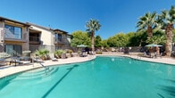Located at 5757 Will Ruth Ave in El Paso, Texas, this outdoor amenity area showcases a generously sized, freeform swimming pool with crystal-clear turquoise water, perfectly suited for the sunny West Texas climate. The pool deck is finished with smooth concrete and is lined with lounge chairs for sunbathing, as well as shaded patio seating with umbrella-covered tables ideal for relaxing or socializing. A separate hot tub or spa is positioned adjacent to the main pool, offering a stepped entry and a comfortable soaking area. Tall palm trees punctuate the landscape, providing a resort-like atmosphere alongside dense mature trees and desert-adapted plantings that add greenery and natural privacy to the space. A secure perimeter fence encloses the entire area, ensuring safety and a sense of seclusion. The surrounding multi-story residential buildings, featuring balconies and a neutral stucco exterior, frame the pool area and contribute to the cohesive architectural aesthetic. An American flag is prominently displayed on one of the nearby structures. The space is well-lit with tall pole lighting, making it functional during evening hours. Overall, the outdoor area offers a well-appointed, inviting retreat designed for recreation and relaxation.