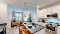This single-story unit in Destin, Florida, offers a thoughtfully designed open-concept layout that maximizes functionality and style. Light wood-look flooring flows throughout, complementing the bright white walls and creating a cohesive, airy atmosphere.

The kitchen is a standout feature, equipped with white shaker cabinets, dark countertops, a subway tile backsplash, and a full suite of stainless steel appliances. A central island with seating serves as both a prep space and casual dining area, illuminated by pendant lighting in mixed metallic finishes. The kitchen opens seamlessly into the living area, which receives ample natural light through large windows fitted with blinds and floor-length curtains. A ceiling fan overhead and a private exterior door add to the room's comfort and practicality.

A built-in desk nook between the kitchen and bedroom provides a dedicated workspace. The bedroom is well-proportioned, featuring a ceiling fan, a natural-light window, and direct access to the full bathroom. The bathroom offers a white vanity with dark countertops, a tub/shower combination with subway tile surround, and in-unit stacked laundry conveniently housed in an adjacent closet.