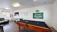 This single-floor recreational facility is designed to serve as a vibrant community hub, offering a variety of spaces for leisure, socializing, and entertainment. The space is unified by consistent white walls, wood-look flooring, and fluorescent lighting throughout.

The centerpiece of the facility is a spacious game room, prominently displaying UVUSA Wolverines branding, including a bold green "Proud Wolverines" banner and the team's mascot logo. This area is home to a handsome wooden pool table with a black felt surface and a foosball table, providing ample entertainment options. A wall-mounted flat-screen television adds another dimension to the room, making it ideal for watching games or events.

Adjacent to the game room is a comfortable lounge area furnished with a large, dark leather sectional sofa. Two windows allow natural light to filter in, offering views of greenery outside. A wall-mounted mini-split air conditioning unit ensures year-round comfort, while additional seating and wall-mounted equipment add functional character to the space.

A separate dining and refreshment area rounds out the facility, featuring multiple round high-top tables with stools, a countertop bar ledge, and a compact refrigerator — perfect for casual gatherings. Windows throughout the space maintain a bright, open atmosphere. Overall, the space is well-suited for group recreation, community events, and relaxed socializing.