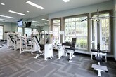 This fitness center offers a thoroughly outfitted workout environment designed to accommodate a wide range of exercise routines. The space is thoughtfully laid out across a single open floor, with dedicated zones for both cardiovascular training and strength equipment. Along one side, a row of treadmills and elliptical machines provides ample options for cardio workouts, while a robust collection of cable machines, weight stations, and resistance equipment lines the opposite side. A full-length mirror spans one wall, enhancing the sense of space and allowing users to monitor their form during exercise. Mounted flat-screen televisions are positioned throughout for convenience during workouts. Natural light pours in through multiple large, multi-pane windows on several walls, offering views of the surrounding outdoor areas and creating a bright, welcoming atmosphere. Ceiling fans complement the recessed and panel lighting overhead, ensuring comfortable air circulation throughout. The flooring features durable, patterned carpet tiles that define workout zones and provide practical underfoot support. A dumbbell rack and free weight area round out the equipment offerings, making this a comprehensive facility suitable for a full-body fitness routine.