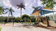 This outdoor playground is designed to provide a well-equipped recreational space within a tropical setting. The play area is surfaced with light-colored gravel, offering a safe and durable ground cover throughout. A central multi-unit play structure serves as the focal point, featuring slides, climbing walls, and elevated platforms, all sheltered by large shade sails that provide meaningful protection from direct sun exposure. Alongside this main structure, freestanding play elements include a spinner and climbing apparatus, adding variety to the activity options available.

The playground is fully enclosed by a dark metal perimeter fence, enhancing safety while maintaining clear sightlines to the surrounding landscape. Tall royal palms rise prominently above the space, complemented by a dense backdrop of mature tropical trees and shrubs that create a lush, green boundary. The overall setting benefits from open sky exposure, allowing natural light to fill the area throughout the day. The combination of structured play equipment, thoughtful shade provisions, secure fencing, and mature tropical vegetation gives this playground a well-maintained and purposefully designed character suited to an outdoor recreational environment.