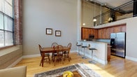 This two-story loft-style residence showcases an industrial-inspired design, blending raw architectural elements with modern finishes throughout. The main level greets you with soaring ceilings that amplify the open-concept layout, where the living, dining, and kitchen areas flow seamlessly together. Light hardwood flooring runs throughout, complementing warm-toned walls and generous windows—including large panes framed by exposed brick—that invite abundant natural light and outdoor views.

The kitchen is well-appointed with rich wood cabinetry, granite countertops, stainless steel appliances, and a raised breakfast bar with bar seating. The adjacent dining area comfortably accommodates a full table, while the living space offers a relaxed setting beneath an open view to the upper loft above, framed by a metal railing.

Two bedrooms on the lower level feature hardwood floors and closet access, while the shared bathroom includes a granite-topped vanity, tile flooring, and an in-unit washer/dryer. The upper loft level, accessed via staircase, provides additional flexible living space with carpeted floors, exposed ductwork, and room for a lounge area and home office.