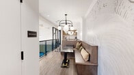 Located at 2025 Cleveland Ave in Charlotte, NC, this thoughtfully curated space offers two distinct dining areas, each with its own character and purpose. The first dining area exudes a warm, inviting ambiance centered around a built-in wood banquette lined with an assortment of textured throw pillows. A striking marble-topped table anchors the seating, while an elegant globe chandelier with matte white orbs casts soft, ambient light above. The walls feature an intricate embossed floral wallcovering in muted neutral tones, adding an artistic, dimensional quality to the space. Warm wood-look flooring and crown molding details lend a refined, polished finish, while views of an outdoor area with a pool are visible through black-framed glass railings nearby. The second dining area offers a more intimate, meeting-oriented atmosphere with a solid wood table surrounded by tufted leather chairs set atop a richly patterned carpet in deep blue and grey tones. The dark-painted ceiling with decorative crown molding creates a dramatic contrast against the crisp white wainscoted walls. A wall-mounted display screen is flanked by an eye-catching arrangement of decorative brass key sculptures, adding a distinctive artistic focal point. An ornate chandelier completes the refined, upscale aesthetic of this versatile dining environment.