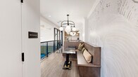 Located at 2025 Cleveland Ave in Charlotte, NC, this thoughtfully curated space offers two distinct dining areas, each with its own character and purpose. The first dining area exudes a warm, inviting ambiance centered around a built-in wood banquette lined with an assortment of textured throw pillows. A striking marble-topped table anchors the seating, while an elegant globe chandelier with matte white orbs casts soft, ambient light above. The walls feature an intricate embossed floral wallcovering in muted neutral tones, adding an artistic, dimensional quality to the space. Warm wood-look flooring and crown molding details lend a refined, polished finish, while views of an outdoor area with a pool are visible through black-framed glass railings nearby. The second dining area offers a more intimate, meeting-oriented atmosphere with a solid wood table surrounded by tufted leather chairs set atop a richly patterned carpet in deep blue and grey tones. The dark-painted ceiling with decorative crown molding creates a dramatic contrast against the crisp white wainscoted walls. A wall-mounted display screen is flanked by an eye-catching arrangement of decorative brass key sculptures, adding a distinctive artistic focal point. An ornate chandelier completes the refined, upscale aesthetic of this versatile dining environment.