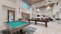 This single-floor amenity and social space is defined by its dramatic double-height ceilings, which lend an expansive, airy quality to the entire area. A warm wood-paneled ceiling accent contrasts beautifully with the crisp white walls, while a series of large-format tile floors laid in a geometric diamond pattern anchors the open layout with visual sophistication.

The main gathering area serves as the social hub, featuring a full-size pool table with a green felt surface and a substantial shuffleboard table, both crafted in rich dark wood. Overhead, multiple circular chandelier fixtures provide layered ambient lighting. A central high-top table with gold-toned legs and white marble surface offers additional seating.

Floor-to-ceiling arched windows and sets of French doors line one wall, flooding the interior with natural light and offering views of surrounding greenery. An ornate wrought-iron gate adds an architectural focal point near the entry corridor, which connects to adjacent hallways via elegant arched openings.

A dedicated game room nook features a round card table surrounded by upholstered chairs, a wall-mounted television, and sheer drapery softening the natural light. A lounge area with club chairs and additional televisions completes the social amenities, creating a versatile and well-appointed communal environment.