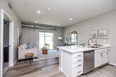 This thoughtfully designed one-bedroom, one-bathroom unit offers a contemporary living experience across a single floor. A welcoming entry hallway with light wood-style flooring leads into the open-concept main living area, where the kitchen and living room flow seamlessly together. The kitchen is outfitted with crisp white shaker cabinetry, light quartz countertops, stainless steel appliances including a range and microwave, and a sink with ample counter space on both sides. An architecturally distinctive feature throughout is the exposed metallic ductwork running overhead, lending the space an industrial-modern character that pairs well with the neutral palette and clean finishes. The living room is bright and inviting, anchored by large sliding glass doors that flood the space with natural light and provide outdoor views. The bedroom offers a cozy retreat with plush carpeting, a generous window with abundant natural light, and a ceiling fan. It connects directly to a spacious walk-in closet with wire shelving on multiple walls. The full bathroom features a tub-shower combination, white vanity cabinetry, and wood-style flooring. A dedicated laundry room with a full-size washer and dryer adds everyday convenience, completing this well-appointed and efficiently organized living space.