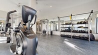 Located at 644 Chlois Ln in Columbus, Ohio, this fitness center is a thoughtfully designed, single-level facility offering a comprehensive range of exercise equipment in a bright, modern setting. The space is divided into distinct workout zones that together accommodate a full spectrum of training needs.

The cardio area is positioned along a wall of large, floor-to-ceiling windows that flood the space with natural light and provide views of the surrounding urban environment. Life Fitness treadmills and elliptical trainers are arranged in this section, each equipped with personal display screens. Ceiling-mounted televisions are positioned throughout for added entertainment during workouts.

The strength and free weights area features a well-stocked dumbbell and kettlebell rack, a TRX suspension training station, and a multi-functional cable machine. Full-length mirrors line the walls, enhancing the sense of openness while providing functional feedback during exercises. Adjustable benches are distributed throughout for versatile use.

A dedicated cycling area includes multiple stationary spin bikes, positioned near additional large windows that invite natural light and outdoor views. The flooring throughout is a durable, textured rubber surface suited for heavy equipment use. Recessed lighting and ceiling fans complement the natural light, maintaining a clean, contemporary ambiance across the entire facility.