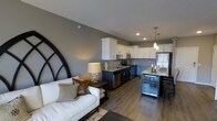 This single-floor space showcases a thoughtfully designed open-concept layout that seamlessly connects the living area and kitchen. The living space features warm gray walls, recessed lighting, and durable wood-look flooring that flows throughout, creating a cohesive and inviting atmosphere.

The kitchen is a standout feature, outfitted with crisp white upper cabinetry and dark lower cabinetry that create a striking two-tone contrast. Light granite countertops provide ample workspace, while stainless steel appliances — including a refrigerator, range, microwave, and dishwasher — add a sleek, modern touch. Elegant pendant lights hang above a peninsula that doubles as a dining surface, and a built-in wine cooler positioned at the end of the island adds a sophisticated entertaining element.

The adjoining living area is anchored by a white sofa accented with textured throw pillows, complemented by a bold arched decorative mirror that adds architectural character to the gray accent wall. A warm-toned table lamp provides ambient lighting, enhancing the cozy yet contemporary feel of the space. Together, the rooms offer a well-balanced blend of functionality and modern style, ideal for both everyday living and entertaining.