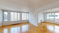 This single-floor residence is characterized by its warm parquet hardwood flooring, which extends consistently throughout the living areas, dining space, and bedrooms, lending a cohesive and polished aesthetic to the interior. The layout is thoughtfully organized, with a spacious living room anchored by large windows that allow abundant natural light to fill the space while offering views of the surrounding urban environment. An adjoining dining area features a classic chandelier overhead and access to additional windows, reinforcing the bright, airy atmosphere.

The galley-style kitchen is outfitted with white cabinetry, granite-look countertops, and white appliances including a gas range, built-in microwave, dishwasher, and refrigerator. Light ceramic tile flooring distinguishes the kitchen from the rest of the unit. A corridor connects the main living areas to the bedroom, which benefits from its own set of large windows and a closet. A dedicated walk-in storage closet provides substantial shelving and hanging space. The full bathroom includes a tiled tub-shower combination with clean, neutral finishes. Throughout the unit, white walls and trim maintain a clean, functional presentation.