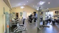 This fitness center is a thoughtfully designed workout facility that provides a comprehensive range of exercise equipment for both strength training and cardiovascular fitness. The space is defined by its vaulted ceilings, which lend an open, airy feel to the room and contribute to a comfortable workout atmosphere. Recessed lighting and ceiling fans are evenly distributed throughout, ensuring adequate illumination and airflow during exercise sessions.

The floor is finished with durable rubber flooring, a practical choice that provides safety and sound absorption. A wide selection of strength machines lines the space, including leg press equipment, cable machines, and various resistance stations designed to target multiple muscle groups. Cardio equipment is also present, rounding out the facility's offerings for a full-body fitness experience.

Large mirrors are mounted along one wall, allowing users to monitor their form during workouts and also visually expanding the sense of space within the room. A wall-mounted television provides entertainment options during exercise. The warm yellow walls and clean white trim give the facility a welcoming, well-maintained appearance. An adjacent room is accessible through a doorway, suggesting additional support amenities nearby. Overall, this fitness center is a functional and inviting space suited for a broad range of workout routines.