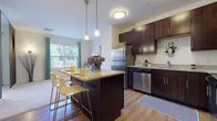 This single-floor residence offers a thoughtfully designed open layout that seamlessly connects the kitchen and living areas. The kitchen is a standout feature, showcasing rich dark-wood cabinetry with sleek hardware, granite countertops, and a full suite of stainless steel appliances including a refrigerator, dishwasher, microwave, and range. A central island with a granite surface provides additional prep space and doubles as a casual dining area with bar-height seating, all illuminated by stylish pendant lighting. Light wood-style flooring runs throughout the kitchen and extends into connecting areas, while plush carpeting defines the living and bedroom spaces.

The living area is bright and open, featuring neutral walls and a clean, minimalist aesthetic that invites personalization. Large windows in the bedroom frame lush outdoor greenery, allowing generous natural light to fill the room. A sleek sliding panel divider adds a modern architectural touch to the bedroom space. The bathroom is well-finished, featuring a tub-shower combination with tile surround, wood-style flooring, a dark-cabineted vanity, and a crisp white countertop. A convenient in-unit stacked washer and dryer rounds out this functional and stylish living space.