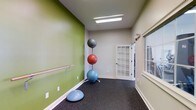This single-floor fitness studio is thoughtfully designed to support a variety of exercise activities. The room features durable dark rubber flooring, which provides cushioning and traction ideal for workouts, stretching, and balance training. A wall-mounted ballet barre runs along the bold olive-green accent wall, making the space suitable for dance, yoga, or stretching routines. The remaining walls are finished in a neutral light gray, creating a clean contrast with the vibrant green feature wall.

The studio is equipped with a vertical stability ball rack holding multiple exercise balls of varying sizes, along with a BOSU balance trainer on the floor, offering practical fitness tools readily available within the space. Overhead, a flush rectangular fluorescent light fixture provides bright, even illumination throughout the room.

A classic white-framed glass-paned door leads to an adjacent area, while a large window along one wall invites natural light into the space, enhancing the open and energetic ambiance. White baseboards throughout add a crisp, finished look. Overall, the studio strikes a balance between functional fitness design and a clean, welcoming aesthetic.
