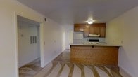 This single-story space presents an open-concept layout that integrates the kitchen and living area into one cohesive environment. The kitchen is equipped with a suite of white appliances, including a refrigerator and electric range, complemented by a built-in range hood. Warm-toned wooden cabinetry lines the upper walls, providing ample storage, while a peninsula with a light-colored countertop serves as a natural divider between the kitchen and the open living space. A flush-mount ceiling light illuminates the kitchen area. The living space features neutral-toned walls and a textured ceiling, contributing to a practical, understated aesthetic. A doorway on the left side of the room leads to an adjacent space, hinted at by the presence of louvered closet doors, suggesting a bedroom or utility area. The flooring in the main area appears to be in a transitional state, with subfloor visible. A door at the far end of the space allows for natural light and suggests an additional exit or entry point. Overhead ventilation is present throughout, supporting climate control across the space.