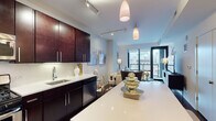 This stylish single-floor urban residence offers a thoughtfully designed open-concept layout that seamlessly connects the kitchen, dining, and living areas. The kitchen is a standout feature, showcasing rich dark-wood cabinetry, gleaming white quartz countertops, a large center island, stainless steel appliances including a gas range, built-in microwave, and French door refrigerator, as well as a classic subway tile backsplash and hardwood flooring. Pendant and track lighting throughout the kitchen and living area create a warm, contemporary ambiance. The living room is an inviting retreat with neutral-toned walls, stylish décor accents, and direct sightlines to expansive floor-to-ceiling windows that frame impressive city skyline views. The bedrooms continue the refined aesthetic, featuring plush carpeting, large windows with striking urban vistas, and carefully considered design details including statement mirrors and layered textiles. The bathroom is clean and bright, finished with large-format tile flooring, wall-mounted fixtures, and curated artwork. Throughout the residence, the interplay of natural light, modern finishes, and cohesive design elements creates a polished, comfortable living environment ideally suited to urban life.