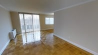 This single-floor space showcases a bright, open living area finished with glossy parquet-style block flooring that adds warmth and visual interest underfoot. The room benefits from generous natural light streaming in through large sliding glass doors fitted with vertical blinds, which open to an outdoor balcony or patio area, providing a seamless connection to the exterior. A secondary window on the adjacent wall further enhances the light-filled atmosphere, offering additional outdoor views. The walls are painted in a neutral warm beige tone, complemented by crisp white baseboards and clean crown moulding that frames the ceiling, lending the space a polished, well-finished appearance. The open layout creates a versatile living environment with a sense of spaciousness, suitable for a variety of furniture arrangements. The overall ambiance is clean, bright, and functional, with the reflective quality of the flooring amplifying the natural light throughout the room.