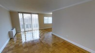This single-floor space showcases a bright, open living area finished with glossy parquet-style block flooring that adds warmth and visual interest underfoot. The room benefits from generous natural light streaming in through large sliding glass doors fitted with vertical blinds, which open to an outdoor balcony or patio area, providing a seamless connection to the exterior. A secondary window on the adjacent wall further enhances the light-filled atmosphere, offering additional outdoor views. The walls are painted in a neutral warm beige tone, complemented by crisp white baseboards and clean crown moulding that frames the ceiling, lending the space a polished, well-finished appearance. The open layout creates a versatile living environment with a sense of spaciousness, suitable for a variety of furniture arrangements. The overall ambiance is clean, bright, and functional, with the reflective quality of the flooring amplifying the natural light throughout the room.