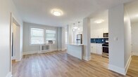 This thoughtfully renovated single-floor residence combines modern finishes with a clean, bright aesthetic throughout. The open-concept living and kitchen area serves as the heart of the space, where warm light wood flooring flows seamlessly underfoot and large windows dressed with blinds flood the room with natural light. A radiator beneath the windows provides classic in-unit heating, while a window-mounted AC unit adds year-round climate control.

The kitchen is a standout feature, designed with crisp white cabinetry, gleaming quartz countertops, and a striking blue herringbone tile backsplash that adds a bold design element. A peninsula with pendant lighting above creates a natural divide between the kitchen and living area. Stainless steel appliances, including a refrigerator, gas range, microwave, and dishwasher, complete the fully equipped space.

The bedroom is a calm and airy retreat, featuring the same light wood flooring, soft neutral walls, and generous window coverage that brings in ample natural light. The bathroom is elegantly finished with large-format stone-look floor tiles and a full soaking tub surrounded by dramatic deep navy herringbone wall tile with brushed gold hardware. A hallway nook houses a stacked washer and dryer, offering the convenience of in-unit laundry.