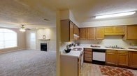 This single-story space is designed with functionality and comfort in mind. The kitchen is well-equipped, featuring an L-shaped layout with ample warm-toned wooden cabinetry that provides generous storage both above and below the counter. White laminate countertops offer a clean, contrasting surface, while the space accommodates key appliances including an electric range and a dishwasher. Under-cabinet lighting adds practicality to the workspace. The kitchen transitions seamlessly into the adjoining living area, which is finished with plush carpeting that adds warmth underfoot. A fireplace serves as a focal point in the living area, framed by a simple white mantle, creating an inviting atmosphere. Natural light filters into the living space through a window near the entry door, brightening the neutral-toned walls. The flooring transitions from carpet in the living area to hard surface in the kitchen, combining practicality with comfort. The textured ceiling and recessed ventilation throughout reflect a thoughtful and efficient use of the overall layout, making this a well-rounded and livable space.