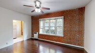 This one-bedroom, one-bathroom unit blends classic character with functional design across a single floor. The living room serves as the centerpiece of the space, showcasing a striking exposed red brick accent wall, rich dark hardwood flooring, and a ceiling fan with light fixture. A multi-pane white-framed window allows natural light to filter in, offering views of surrounding greenery and creating a warm, inviting atmosphere. A wall-mounted heater adds a practical touch to the room.

The kitchen features rich cherry wood cabinetry, granite-look countertops, a stainless steel double sink, and a full-size stainless steel refrigerator, all set atop light tile flooring. The layout is compact yet functional, offering ample storage.

The bedroom continues the hardwood flooring theme and is illuminated by a ceiling fan and a window. A closet door provides convenient built-in storage, and the clean white walls keep the room bright and versatile.

The bathroom is finished with white subway tiles accented by a decorative mosaic border, a pedestal sink, a soaking tub with overhead shower, and light tile flooring — delivering a clean, timeless aesthetic. A hallway connects the rooms, completing this well-organized and character-rich unit.