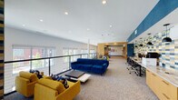 This thoughtfully designed single-floor amenity space blends collaborative work environments with comfortable social areas, all unified by a contemporary aesthetic. The open lounge area anchors the space with plush navy and mustard-yellow seating arranged around low-profile coffee tables, set against a backdrop of expansive windows fitted with roller shades. A sleek cable-wire railing runs along the window-facing perimeter, framing panoramic outdoor views of the surrounding neighborhood.

A tiled accent column serves as a bold architectural focal point, clad in a geometric pattern of blue, gold, and neutral tones that adds visual interest throughout. Warm wood-paneled wall treatments define transitional zones, lending a refined contrast to the crisp white ceilings lined with recessed lighting.

The co-working zone features individual workstations along a navy accent wall, while two enclosed conference rooms offer privacy for meetings. Each conference room is equipped with a wall-mounted display, a rich wood conference table, ergonomic seating, and distinctive patterned wallcoverings. A banquette-style lounge nook with navy upholstery, round wood tables, and pendant sconce lighting provides a more intimate setting. Curated artwork and built-in desk nooks complete the space.