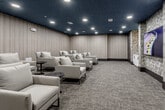Located at 13220 Foster St in Overland Park, Kansas, this refined screening and lounge space offers a thoughtfully designed environment built for comfort and entertainment. The room is defined by its striking deep navy ceiling, which creates an immersive, theater-like atmosphere enhanced by recessed can lighting arranged to provide even, ambient illumination throughout.

The walls are clad in warm, linear wood-paneled panels that lend a contemporary and sophisticated aesthetic, complemented by sections of bold geometric patterned wallcovering that add visual interest and depth. Plush, light-toned upholstered lounge chairs are arranged in a tiered, screening-room fashion across textured, multi-tone carpeting, offering generous seating for groups. Sleek dark round side tables are positioned throughout for added convenience.

A dedicated media area features a wall-mounted flat-screen television set against the geometric accent wall, flanked by stylish sconce lighting and a white cabinetry unit with a marble-look countertop, ideal for refreshments or equipment. The entryway area continues the design language with matching finishes and additional storage cabinetry. Built-in speakers are integrated into the walls, underscoring the room's commitment to a full audio-visual experience. The overall ambiance is intimate, polished, and purpose-built for private screenings or curated gatherings.