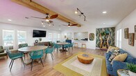 This charming clubhouse in Memphis, TN, offers a welcoming, open-concept layout that seamlessly connects the living room, dining area, and kitchen into one cohesive, functional space. The interior is anchored by warm wood-look flooring throughout and accented by striking tropical-themed wallpaper murals that lend the space a bold, resort-inspired personality.

The living area features a plush blue sofa, a rustic wood drum coffee table, and a yellow geometric area rug that adds a pop of color. Natural light pours in through multiple windows with white shutters, while a decorative ladder shelf styled with plants and accessories adds character near the entry. Exposed wood ceiling beams and dual ceiling fans, complemented by adjustable track lighting, give the space an airy, relaxed feel.

The dining zone accommodates multiple round tables surrounded by vibrant teal lattice chairs, with a wall-mounted flat-screen TV and a warm wood sideboard creating a comfortable social hub.

The kitchen is a standout feature, showcasing bold navy blue cabinetry with gold hardware, white countertops, and a dramatic circular-patterned accent wall. Stainless steel appliances — including a range, dishwasher, and refrigerator — make this kitchen fully functional for events and gatherings. A pass-through window connects the kitchen to the main space, enhancing the open, communal flow of the entire clubhouse.