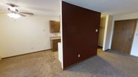 This single-floor space is defined by a striking dark red dividing wall that serves as the central architectural feature, visually separating the carpeted living area from the kitchen zone. The living area features neutral, wall-to-wall carpet in a warm beige-gray tone, providing a grounded, understated base throughout the space. The dividing wall is finished in a deep burgundy red, creating a bold contrast against the white surrounding walls and adding a distinct focal point to the layout.

The kitchen area, partially visible beyond the divider, features warm oak cabinetry with a classic raised-panel design. A countertop workspace with lower cabinetry provides functional prep and storage space. The kitchen transitions seamlessly into the adjacent area via an open pass-through design at the base of the dividing wall.

A textured ceiling runs throughout the space, consistent with the overall construction style. To the right side, a closet area is visible, suggesting additional storage capacity. The space is illuminated by overhead lighting and benefits from a straightforward, functional layout suited for everyday living.