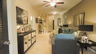 This single-floor residence offers a cohesive and thoughtfully designed living environment that balances comfort with style. The main living area is anchored by a plush, wall-to-wall carpet that adds warmth underfoot, while a ceiling fan with integrated lighting provides both function and a subtle focal point overhead. The space is bathed in a soft, ambient glow that highlights the neutral, beige-toned walls — a versatile backdrop that complements a range of furnishings and décor. A comfortable seating area with a blue sofa and accent pillows invites relaxation, while a sleek mirrored console table along the wall adds a touch of elegance and visual depth. Curated artwork, including a striking equestrian piece, contributes character to the living space. Moving deeper into the layout, a dining nook with bar-height seating transitions naturally into what appears to be a well-appointed kitchen area, complete with countertop workspace and cabinetry visible in the background. The open sightlines between the living, dining, and kitchen zones create a sense of spaciousness and ease of flow throughout the home. Overall, the space presents a warm, inviting ambiance suited for both everyday living and casual entertaining.