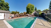 Located at 11805 Dry Creek Rd in Auburn, California, this outdoor community pool area offers a well-maintained and inviting recreational space designed for leisure and relaxation. The centerpiece is a generously sized rectangular swimming pool with clear, blue-green water, framed by a clean concrete pool deck that provides ample space for lounging and movement around the perimeter.

The pool area is thoughtfully equipped with multiple patio umbrella stations, each accompanied by seating, offering shaded retreats from the warm Northern California sun. A dedicated utility structure on the pool deck houses essential pool equipment and safety supplies, including a clearly posted fire extinguisher, contributing to the area's safety-conscious design.

The space is enclosed by a perimeter fence, ensuring privacy and security, while mature trees — including tall conifers and broad deciduous varieties — line the surroundings, creating a natural, park-like atmosphere. Seasonal color from flowering shrubs adds visual interest to the landscape backdrop.

Neighboring residential buildings with wood-paneled exteriors are visible, integrating the pool area naturally into the broader community setting. Under clear blue skies, the space enjoys abundant natural light throughout the day, making it an ideal communal outdoor amenity for residents seeking both active recreation and relaxed outdoor enjoyment.