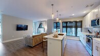 Located at 205 S High St in the heart of downtown Columbus, Ohio, this modern single-floor unit offers a thoughtfully designed open-concept layout. The combined kitchen and living area serves as the centerpiece of the space, flooded with natural light through large floor-to-ceiling windows that frame views of the surrounding cityscape. Light hardwood flooring runs throughout, complementing the neutral gray walls and bright white trim.

The kitchen is well-appointed with crisp white shaker cabinetry extending to the ceiling, providing ample storage. A quartz-topped island with bar seating and elegant globe pendant lights anchors the space, while stainless steel appliances—including a refrigerator, range, and microwave—complete the functional yet stylish design. A subway tile backsplash adds a clean, contemporary touch.

The living area is open and airy, with wall-mounted television, large windows with draping curtains, and city views that bring an urban energy to the interior.

The bathroom features white shaker vanity cabinetry with a quartz countertop, a full soaking tub with subway tile surround, and a sliding glass enclosure. A dedicated closet provides practical storage. The entrance offers a smooth transition into the main living areas, rounding out a cohesive and well-organized floor plan ideal for urban living.