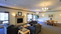 This single-story common lounge area offers a comfortable and well-appointed gathering space designed for relaxation and socialization. The room is anchored by a classic brick fireplace with a wooden mantel, topped with a decorative wall clock and flanked by wall sconces that cast a warm, ambient glow throughout the space. Multiple dark upholstered sofas and armchairs are arranged around glass-topped coffee tables, creating several inviting conversation areas. An elegant chandelier hangs from the textured ceiling, complementing the wall sconces to provide layered lighting across the room. A decorative border trim runs along the upper perimeter of the walls, adding a traditional design element to the space. Natural light filters in through multiple windows dressed with dark curtains, offering views of outdoor greenery. A dining area with a small table and chairs is visible toward the back of the room, adding functional versatility to the space. A large potted plant near the windows contributes a natural, refreshing touch. The combination of soft carpeting, warm lighting, and thoughtfully arranged furnishings creates a cozy and welcoming communal environment.