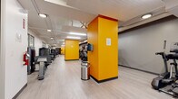 Located at 1965 E Sixth St in Cleveland, Ohio, this single-floor fitness center offers a well-organized and energetic workout environment. The space is defined by its bold orange accent columns, which punctuate the layout and add a vibrant visual identity against the neutral gray walls and light wood-look flooring. Exposed ductwork and overhead fluorescent and recessed lighting illuminate the entire area efficiently, creating a bright and functional atmosphere throughout.

The facility is comprehensively equipped with True-brand cardio machines, including multiple treadmills, ellipticals, and a stair climber, arranged to allow comfortable spacing between users. A rowing machine and stationary bike round out the cardio offerings, while additional strength and functional training equipment is available throughout the space. Wall-mounted flat-screen televisions are positioned in several zones, providing entertainment during workouts.

The elongated floor plan, as seen in the 3D overview, reveals a generous and continuous workout area with distinct equipment zones organized along its length. Stainless steel waste receptacles and sanitation stations are conveniently placed near the equipment. The overall design is clean, modern, and purpose-built for fitness, with an energetic color scheme and efficient use of space that makes it well-suited for a broad range of exercise routines.