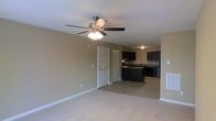 This single-floor space features an open-concept layout that connects the living area and kitchen in one cohesive, flowing environment. The living area is finished with plush carpet and neutral beige walls, creating a warm and inviting atmosphere. A ceiling fan with integrated lighting serves as a central fixture, providing both comfort and illumination to the space. Smooth white trim and baseboards frame the room cleanly throughout. Transitioning seamlessly from the carpeted living area, the kitchen features tile flooring and is outfitted with rich, dark cabinetry that extends along multiple walls, offering generous storage. A centrally positioned kitchen island adds functional workspace and visual definition to the cooking area. Dark stone countertops complement the cabinetry, while a sink is visible along the back wall beneath upper cabinets. The overall color palette — warm beige walls, dark cabinetry, and neutral flooring — creates a balanced contrast that is both practical and aesthetically cohesive. The open flow between living and kitchen spaces makes this layout well-suited for everyday living and entertaining alike. The single-floor design ensures easy accessibility and a comfortable, connected feel throughout the entire space.