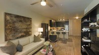 This single-floor space showcases an open-concept layout that connects the living area and kitchen in a cohesive, functional design. The living room is anchored by a comfortable neutral-toned sofa, accented with decorative pillows, and paired with a sleek glass coffee table atop a patterned area rug. A large abstract artwork adorns the wall, adding a bold visual focal point, while a classic table lamp provides warm ambient lighting. A ceiling fan with a built-in light fixture overhead enhances both comfort and airflow.

The kitchen flows naturally from the living area and features dark cabinetry contrasted by a light-colored countertop that doubles as a breakfast bar, complete with modern bar stools. Stainless steel appliances, including a full-sized refrigerator, complement the contemporary aesthetic, while track lighting illuminates the workspace efficiently. A stainless steel sink and carefully arranged countertop accessories reflect a practical yet stylish cooking environment.

Hardwood-style flooring runs throughout the space, unifying the open layout and contributing to a warm, modern ambiance. An interior door visible in the background hints at additional private spaces beyond the main living and kitchen area.