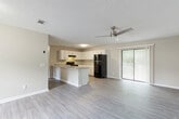 This single-story residence in Pensacola, Florida, presents a clean, modern aesthetic throughout its open-concept layout. Light-toned, wood-look plank flooring runs continuously across the main living areas, lending a cohesive and low-maintenance quality to the space. The living area is generously proportioned, benefiting from a ceiling fan overhead and a sliding glass door fitted with vertical blinds that allows natural light to filter in while providing access to the outdoors. A secondary window further brightens the space.

The kitchen is thoughtfully designed with ample white shaker-style cabinetry extending to the upper walls, offering considerable storage capacity. A peninsula with a neutral-toned countertop defines the boundary between the kitchen and living area, providing additional workspace and a natural gathering point. Black appliances, including a range with an overhead ventilation hood, a refrigerator, and a dishwasher, provide a bold contrast against the white cabinetry. A hallway connects the main living area to the private quarters and bathroom.

The bathroom features a white vanity with a wide mirror, brushed chrome fixtures, a textured ceiling, and a full tub-and-shower combination. A textured ceiling is consistent throughout the entire unit, complementing the neutral wall tones that create a calm, understated atmosphere.
