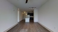 This single-floor residence showcases an open-concept layout that seamlessly connects the living area to the kitchen. The expansive main living space is finished with warm hardwood-style flooring that runs throughout, complementing the clean white walls and creating a bright, welcoming atmosphere. Ceiling-mounted track lighting provides targeted illumination across the living area, while a ceiling fan adds both function and a subtle design element overhead.

The kitchen is positioned toward the rear of the space and is distinguished by sleek dark cabinetry that contrasts sharply with the white central island, which serves as both a prep area and a natural dividing point between the kitchen and living zones. Stainless steel appliances, including a refrigerator and range, add a contemporary touch, while pendant lighting above the island enhances the area's functionality and style. Subway tile backsplash detailing is also visible, contributing to the kitchen's modern aesthetic.

A recessed entryway with a solid door is visible adjacent to the kitchen, offering a defined transition point within the space. Overall, the layout is clean, modern, and efficiently designed.