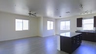 This single-floor space offers a clean, modern aesthetic highlighted by an open-concept layout that seamlessly connects the kitchen and living areas. The kitchen features a sleek, dark-cabineted island topped with polished granite countertops, providing both a functional workspace and a natural visual anchor for the space. Hardwood-style flooring extends throughout, adding warmth and continuity to the interior. The living area is bathed in natural light, courtesy of multiple sliding and casement windows positioned across the surrounding walls, creating a bright and airy atmosphere. A ceiling fan is centrally installed, enhancing comfort and air circulation. The neutral color palette — from the crisp white walls to the light-toned floors — lends the space a versatile, move-in-ready quality. The open floor plan allows for flexible furniture arrangements, making the space well-suited for a variety of living configurations. Overall, the space balances contemporary finishes with a welcoming, practical layout designed to maximize both comfort and functionality.