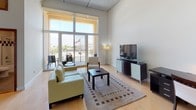 Located at 550 Seagaze in Oceanside, Unit 13 is a contemporary single-floor residence defined by dramatic soaring ceilings and an abundance of natural light. The open-concept living area is anchored by expansive floor-to-ceiling windows and glass doors that frame sweeping views of the surrounding cityscape and glimpses of the coast, flooding the interior with sunlight throughout the day. Light hardwood flooring runs consistently throughout, reinforcing the bright, airy atmosphere.

The kitchen is a standout feature, outfitted with sleek gray cabinetry, stainless steel appliances including a full-size refrigerator, dishwasher, and built-in oven, as well as a quartz-topped island with seating. A distinctive slatted wood ceiling accent adds a warm architectural detail to the kitchen zone. The living area provides generous space with direct access to a private balcony, ideal for taking in the outdoor views.

The bedroom is a serene retreat, featuring towering windows on two walls that capture panoramic city views and fill the room with natural light. Built-in dark wood wardrobes offer ample storage. The bathroom is clean and well-appointed, with a quartz vanity, tile flooring, and a glass-enclosed shower-tub combination accented with decorative tile detailing.
