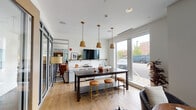 Located on Ellinwood Street in Des Plaines, Illinois, this single-story amenity space is designed with a contemporary aesthetic that emphasizes openness, natural light, and versatile functionality. Light hardwood flooring runs throughout, complementing a neutral white and grey palette accented by warm wood tones and brass hardware details.

The central gathering area features a substantial dark wood communal table with leather-topped brass stools, positioned beneath a trio of pendant lights with brass fittings. A wall-mounted television, open shelving, and a kitchenette with a sink and counter space make this zone well-suited for both casual socializing and organized gatherings. Large floor-to-ceiling windows and sliding glass doors frame views of the surrounding streetscape, flooding the interior with natural light.

Adjacent to this hub, a lounge area offers intimate seating with rounded armchairs and low circular tables arranged near oversized windows. Glass-and-steel partition walls with natural wood-framed doors divide the space into distinct zones while maintaining visual connectivity throughout. A dedicated game area includes a foosball table and curated wall art, adding a recreational dimension. A smaller workstation nook with quartz countertops and cabinetry provides a quieter retreat within the broader space.