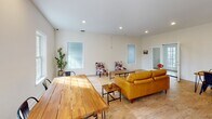 This single-floor space offers an open-concept layout that seamlessly connects the living, dining, and kitchen areas, creating a cohesive and functional environment. The living area is generously sized, featuring light hardwood flooring throughout, recessed lighting, and multiple windows that invite abundant natural light. The neutral white walls and clean lines lend a bright, airy feel to the entire space. Seating in the living area includes a warm caramel leather sofa complemented by distinctive cowhide-print accent chairs arranged around a wooden coffee table and a woven area rug, adding texture and character to the room. The dining area accommodates a solid wood plank table with industrial-style metal chairs and a decorative table runner, positioned near a window with views of lush greenery outside. The kitchen is well-equipped and stylishly designed, featuring crisp white cabinetry, light granite countertops, and stainless steel appliances including a French door refrigerator and built-in microwave. A peninsula with bar seating provides additional prep and casual dining space. Throughout the space, the consistent hardwood flooring, recessed lighting, and clean white palette create a cohesive, modern aesthetic that is both welcoming and practical.