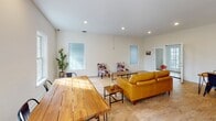 This single-floor space offers an open-concept layout that seamlessly connects the living, dining, and kitchen areas, creating a cohesive and functional environment. The living area is generously sized, featuring light hardwood flooring throughout, recessed lighting, and multiple windows that invite abundant natural light. The neutral white walls and clean lines lend a bright, airy feel to the entire space. Seating in the living area includes a warm caramel leather sofa complemented by distinctive cowhide-print accent chairs arranged around a wooden coffee table and a woven area rug, adding texture and character to the room. The dining area accommodates a solid wood plank table with industrial-style metal chairs and a decorative table runner, positioned near a window with views of lush greenery outside. The kitchen is well-equipped and stylishly designed, featuring crisp white cabinetry, light granite countertops, and stainless steel appliances including a French door refrigerator and built-in microwave. A peninsula with bar seating provides additional prep and casual dining space. Throughout the space, the consistent hardwood flooring, recessed lighting, and clean white palette create a cohesive, modern aesthetic that is both welcoming and practical.