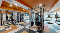 Located at 2801 Windy Ridge Pkwy SE in Smyrna, Georgia, this fitness center offers a spacious, single-floor layout designed to support a comprehensive workout experience. The interior is immediately distinguished by its bold orange-painted ceilings and accent walls, contrasted against crisp white walls and modern recessed lighting, creating an energetic and motivating atmosphere throughout.

The facility is divided into distinct functional zones. The cardio area features a row of treadmills, ellipticals, and a stair climber positioned alongside large floor-to-ceiling windows that flood the space with natural light and provide views of the surrounding outdoor amenities. Overhead ceiling fans enhance comfort across the workout floor.

The strength training zone is outfitted with a robust selection of equipment, including Matrix cable machines, a functional trainer, weight benches, free weights, kettlebells, medicine balls, and plate-loaded machines — catering to a wide range of fitness levels and training styles.

Flooring transitions between a bold multi-toned checkered tile pattern in the main areas and warm wood-look flooring in the free weights and stretching zones, adding visual variety and practical functionality. Wall-mounted televisions are positioned throughout for entertainment during workouts. Glass-panel doors and interior windows connect the zones, maintaining an open and airy feel across the entire facility.