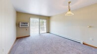 Located at 8800 Macomb St in Grosse Ile, Michigan, this one-bedroom, one-bathroom unit offers a practical and well-organized single-floor layout. The open living area features soft wall-to-wall carpeting, warm neutral tones, and a charming pendant light fixture. A sliding glass door floods the space with natural light and provides access to an outdoor area, while a wall-mounted air conditioning unit ensures year-round comfort. Baseboard heating runs throughout the unit. The kitchen is situated adjacent to the living area in an open-concept arrangement, featuring white cabinetry, white appliances, and hard surface flooring that transitions cleanly from the carpeted living space. The bedroom is a comfortable, carpeted room with a natural wood-trimmed door and closet, a window with outdoor views, and easy access to the full bathroom. The bathroom is clean and functional, appointed with wood-look vinyl flooring, a white vanity with mirror, a standard toilet, and a full tub-shower combination. Warm beige walls and wood-toned trim details carry consistently throughout the unit, creating a cohesive and inviting atmosphere well-suited for comfortable everyday living.
