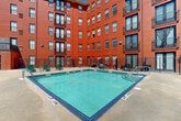 This outdoor amenity space is centered around a generously sized rectangular swimming pool with vivid turquoise tile work, set within a semi-enclosed courtyard formed by the surrounding multi-story red brick building. The architecture is bold and industrial in character, with the warm tones of the brick facade contrasting sharply against clear blue skies. Large windows and private balconies line the building's upper floors, overlooking the pool area below.

The pool deck is finished in concrete and provides ample space for relaxation, lined with green lounge chairs positioned to take advantage of natural sunlight throughout the day. Entry steps with metal handrails offer easy pool access. The courtyard is enclosed by black iron fencing, providing a sense of privacy and security while still maintaining an open, airy atmosphere.

Mature trees visible beyond the perimeter fence add a natural, softening element to the urban setting. Additional amenities near the pool area include a utility station with hose connections and storage. The overall ambiance is lively yet functional, offering residents a well-defined outdoor retreat with a distinctly urban aesthetic defined by the bold red brick architecture that frames the entire space.