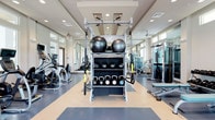This fitness center offers a comprehensive workout environment designed to support a wide range of exercise routines. The space features a two-toned, durable flooring with contrasting light and dark panels that define functional training zones. A central multi-station rack serves as the focal point, stocked with stability balls, medicine balls, kettlebells, and TRX suspension trainers, making it ideal for functional and strength training. Cardio equipment, including ellipticals and treadmills, lines one side of the room, while cable and weight machines occupy the opposite side. Large windows along the perimeter allow ample natural light to fill the space, creating a bright and energizing atmosphere. Mirrored walls enhance the sense of openness and allow users to monitor their form during workouts. Recessed lighting and ceiling fans supplement the natural light, maintaining comfort throughout the space. The clean, modern interior design features neutral tones and white trim, contributing to a professional and well-maintained environment suited for a full-body workout experience.