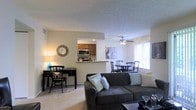 Located at 5450 Firewater Ln in Westerville, Ohio, this single-floor residence offers a well-organized and comfortable layout designed for everyday living. The open-concept living area serves as the heart of the home, featuring plush carpeting, a ceiling fan, and a naturally lit interior enhanced by window blinds that allow for adjustable light control. A charming decorative mirror and tasteful wall art add personality to the space. The living room flows seamlessly into a dedicated workspace area, accommodating a desk and seating — ideal for remote work or study. The kitchen is positioned just beyond a pass-through counter, outfitted with warm wood cabinetry and stainless steel appliances including a microwave and range, making meal preparation both efficient and accessible. Adjacent to the kitchen, the dining area provides ample room for a full dining set, benefiting from natural light streaming through a nearby window. The open sightlines between the living, dining, and kitchen areas create a sense of connected, functional space throughout. The textured ceilings and neutral wall tones contribute to a clean, inviting ambiance that is both practical and welcoming.