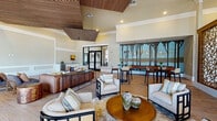 Located at Reed Canal Road in Port Orange, Florida, this single-story clubhouse presents a thoughtfully designed communal space that blends contemporary elegance with warm, natural materials. The interior is immediately distinguished by its soaring ceilings, accented with striking slatted wood ceiling panels that add texture and visual warmth throughout the main gathering areas.

The primary lounge area is generously proportioned, featuring wood-look tile flooring and a curated mix of seating arrangements, including plush upholstered sofas, accent chairs with bold geometric frames, and rustic live-edge coffee tables. A large-scale nature-inspired mural depicting a serene treeline landscape serves as a dramatic focal point along one wall, complementing the bar-height seating area positioned in front of it.

A standout architectural element is the oversized laser-cut decorative wood partition screen, which elegantly defines separate zones within the open floor plan while allowing light and sightlines to pass through. A secondary seating area features additional lounge furniture and French doors that introduce natural light and outdoor views.

The space also includes a practical kitchenette fitted with white shaker cabinetry, granite countertops, a stainless steel sink, and a window overlooking the surrounding landscape. Black-framed glass entry doors complete the polished, resort-style aesthetic.