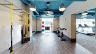 This single-floor fitness studio is thoughtfully designed to support a wide range of workout styles in an energetic and functional environment. The space is immediately distinguished by its bold teal-painted exposed ceiling, which houses visible ductwork and is illuminated by industrial-style pendant lights, creating a modern, gym-boutique aesthetic. The flooring transitions between wood-look vinyl in the main training area and a darker surface in the cycling zone, clearly delineating workout zones.

The main training area is equipped with TRX suspension straps mounted to a wall-mounted bar, a freestanding heavy punching bag, and stacked step platforms and resistance accessories. A large flat-screen display is mounted on the wall, ideal for guided workouts or class instruction. Full-length mirrors framed in teal accents expand the sense of space and support form monitoring during exercise.

An adjoining area houses a row of spin bikes alongside a rack of dumbbells and a colorful collection of stability and medicine balls. A large window in this zone allows natural light to fill the room and offers an outdoor view of surrounding greenery. Multiple gray panel doors provide access to additional rooms, and ceiling fans contribute to comfortable air circulation throughout the space.