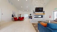 This single-floor community space is designed to impress, combining modern aesthetics with welcoming functionality. Upon entry, a grand reception area greets visitors with soaring vaulted ceilings, large glass-paneled doors, and expansive large-format tile flooring that flows seamlessly throughout the space. A staffed front desk provides a professional focal point near the entrance, accompanied by bold red accent chairs and curated décor that set an inviting tone.

The centerpiece of the main lounge is a striking floor-to-ceiling stone fireplace accented by a natural wood mantel and a wall-mounted television, creating a warm and sophisticated gathering space. Multiple seating arrangements, including plush blue sofas and vibrant red accent chairs positioned atop decorative area rugs, offer versatile options for relaxation and social interaction.

Adjacent to the lounge, a dining area features a sleek round table with upholstered chairs, positioned alongside a well-appointed kitchenette with a marble-topped counter, bar stools, and open wood shelving. This area is bathed in natural light from large windows with views to the exterior.

Throughout the space, vaulted ceilings amplify the sense of openness, while recessed lighting and carefully selected furnishings create a polished, contemporary ambiance suitable for both casual gatherings and everyday community use.